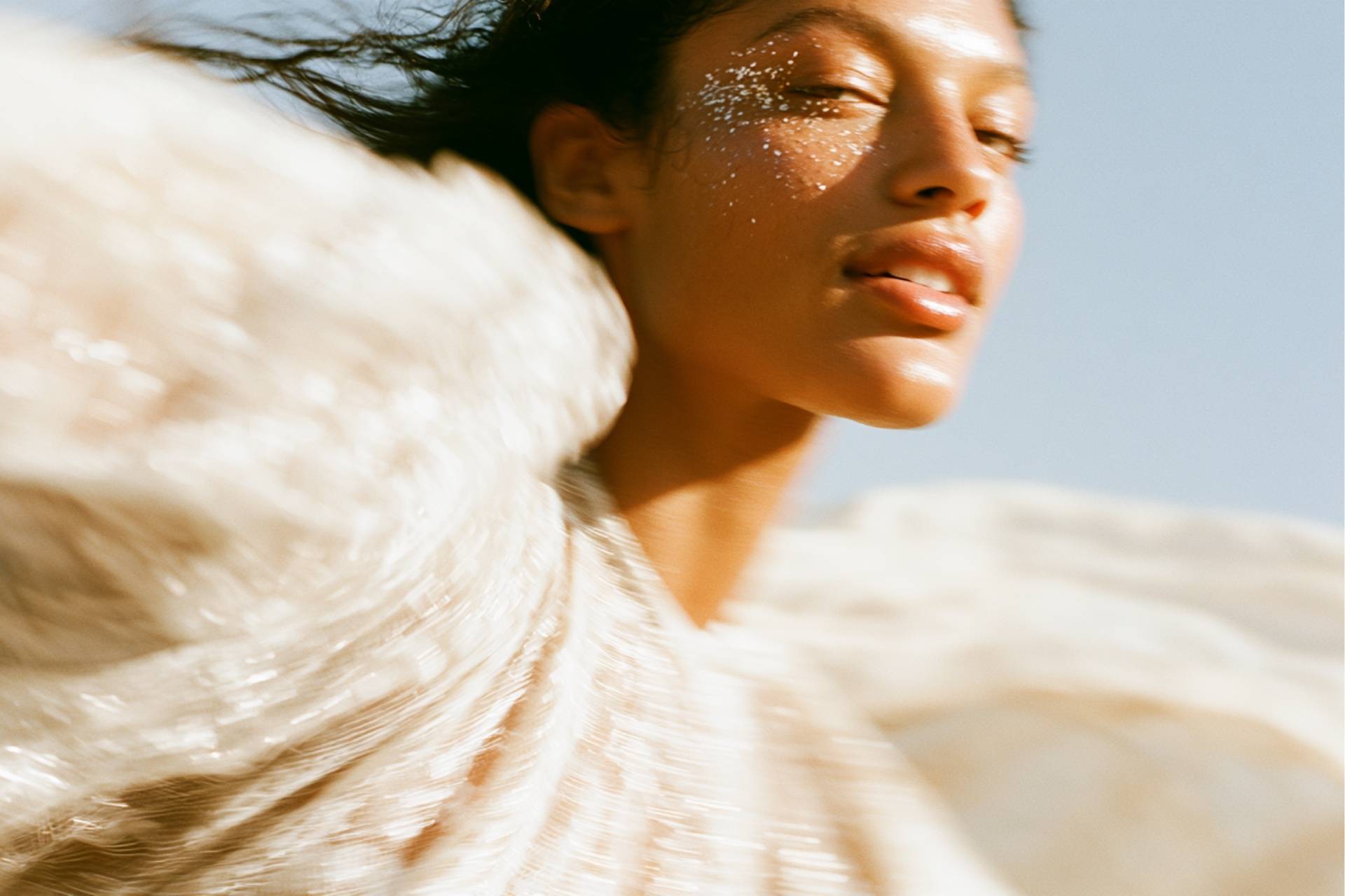 Femme en mouvement sous une lumière solaire franche, paillettes sur la pommette et étoffe crème flottante, univers d'une identité visuelle beauté et bien-être